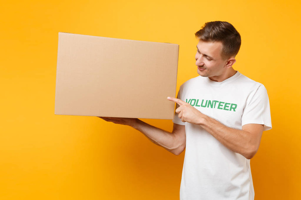 Portrait of good kind man in white t-shirt with written inscription green title volunteer with big cardboard box isolated on yellow background. Voluntary free assistance help, charity grace concept - Photo, Image
