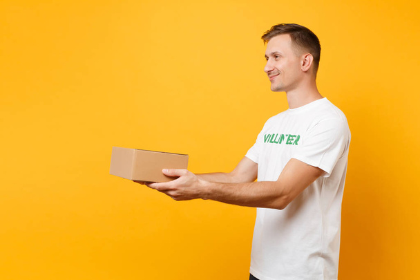 Seitenprofil-Ansicht eines Mannes in weißem T-Shirt mit Schriftzug Grüner Titel Freiwilliger mit leerem Pappkarton auf gelbem Hintergrund. Freiwillige, kostenlose Hilfe, Wohltätigkeitskonzept - Foto, Bild