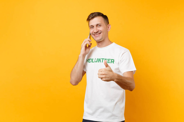 Jonge man portret in wit t-shirt met schriftelijke inscriptie groene titel vrijwilliger praten op mobiele telefoon geïsoleerd op gele achtergrond. Vrijwillige gratis hulp hulp, liefdadigheid gunst werk concept - Foto, afbeelding