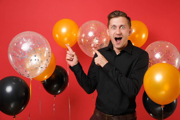 Excited young man in black classic shirt pointing index fingers aside on red background air balloons. St. Valentine's, International Women's Day, Happy New Year, birthday mockup holiday party concept - Photo, Image