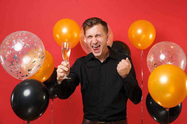 Joven alegre con camisa clásica negra sosteniendo una copa de champán, haciendo un gesto ganador sobre globos aéreos de fondo rojo. Día Internacional de la Mujer Feliz Año Nuevo concepto de fiesta simulada de cumpleaños
 - Foto, Imagen