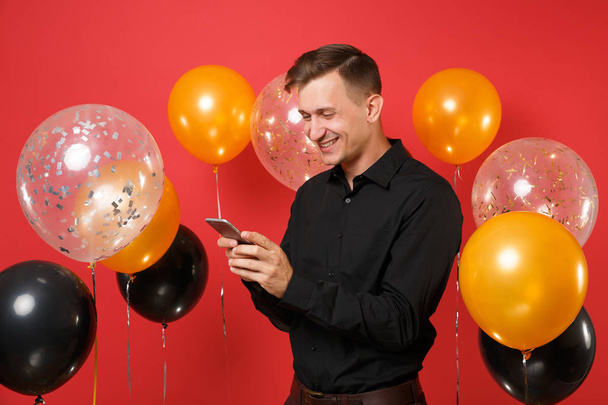 Smiling young man in black classic shirt holding, using mobile phone, typing sms message on red background air balloons. International Women's Day Happy New Year birthday mockup holiday party concept - Photo, Image