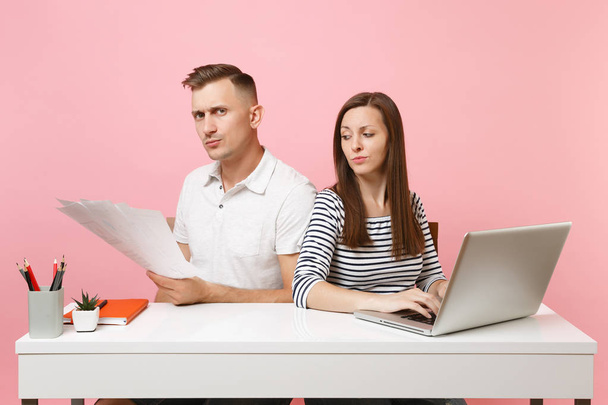 Две молодые сердитые коллеги-деловые женщины сидят за белым столом с современным ноутбуком, изолированным на розовом фоне. Концепция карьерного роста. Копирование космической рекламы, молодежная кооперация
 - Фото, изображение