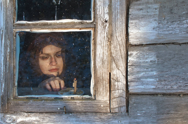 Bäuerin ruht am Fenster in der Nähe einer brennenden Kirchenkerze am Winterabend, vielleicht wartet sie auf jemanden - Foto, Bild