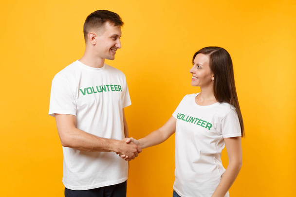 Πορτραίτο του νέους συναδέλφους ζευγάρι σε λευκό t-shirt με γραπτή επιγραφή πράσινο τίτλο εθελοντής απομονώνονται σε κίτρινο φόντο. Εθελοντική εργασία δωρεάν, βοήθεια, φιλανθρωπία χάριτος ομαδική εργασία έννοια - Φωτογραφία, εικόνα