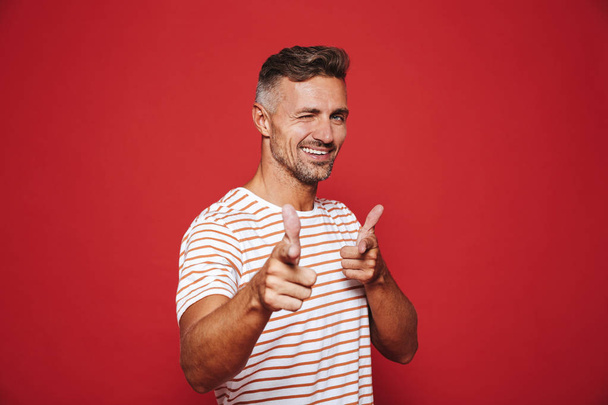 Joyful man in striped t-shirt gesturing index finger on camera meaning hey you isolated over red background - Valokuva, kuva