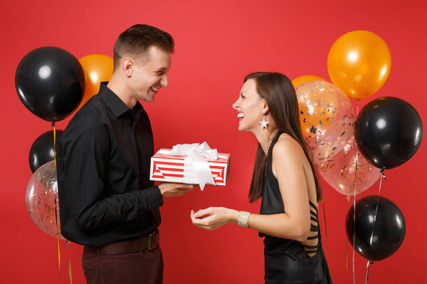 Mladý pár v černých šatech drží dárek box slaví svátek narozenin izolované na jasně červeném pozadí s vrtulovým pohonem St. Valentine International ženy den šťastný nový rok 2019 koncept. - Fotografie, Obrázek