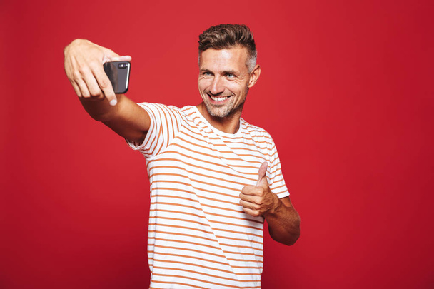 Πορτρέτο του ένας ευτυχισμένος άνθρωπος στέκεται πέρα από το κόκκινο υπόβαθρο, λαμβάνοντας μια selfie - Φωτογραφία, εικόνα