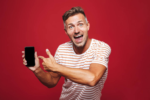 Portrait of an excited man standing over red background, showing blank screen mobile phone, celebrating - Photo, Image