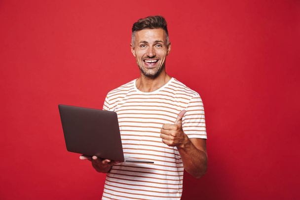 Optimistic unshaved man in striped t-shirt smiling and holding gray laptop isolated over red background - Foto, immagini
