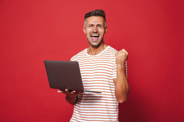 Image of successful guy in striped t-shirt smiling and holding gray laptop isolated over red background - Фото, зображення