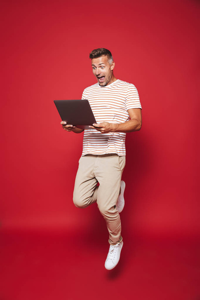 Full length photo of joyful man in striped t-shirt smiling and holding gray laptop isolated over red background - Фото, изображение