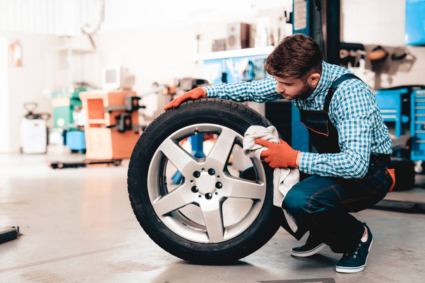 Young Smiling Mechanic Cleans A Car Tire At Service Station. Professional Uniform. Confident Engineer Stare. Cheerful Repairman. Working In The Garage. Protective Gloves. Repair Specialist. - Foto, Imagem