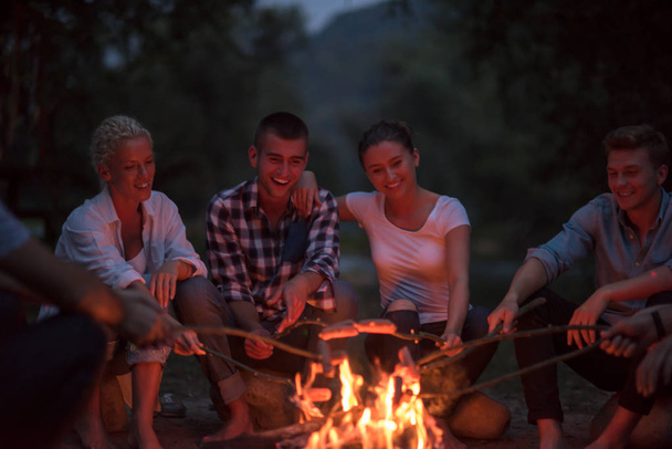 a group of happy young friends relaxing and enjoying  summer evening around campfire on the river bank - Foto, Imagem