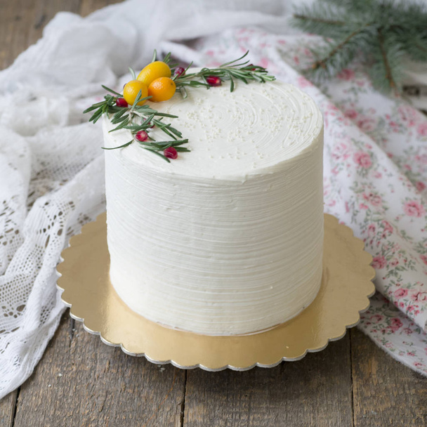 White New Year or Christmas cake, decorated with kumquat, sprigs of rosemary and cranberries on a wooden table. Winter decor. A birthday cake for those born in winter. Copy space, selective focus - Foto, Imagem
