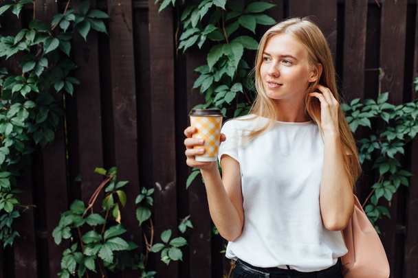 Young beautiful girl with a Cup of coffee, dressed in a white t-shirt, standing in the open air, at the wooden deciduous wall, drinking a drink - Φωτογραφία, εικόνα