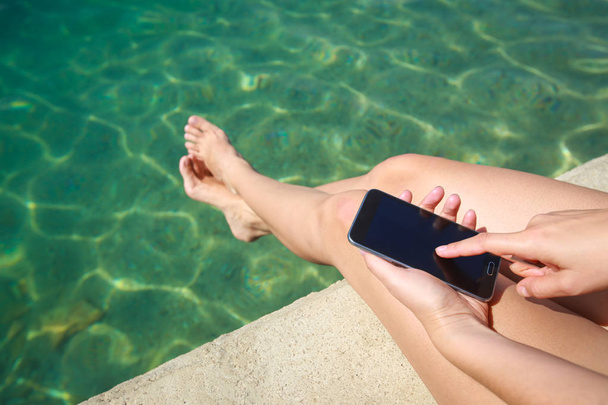 A woman holding smartphone while sitting on the seaside pier. - Valokuva, kuva