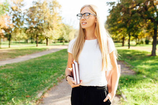 Portrait of a beautiful student girl with glasses, walking in the Park, holding books in her hands, rest after class - Foto, Bild