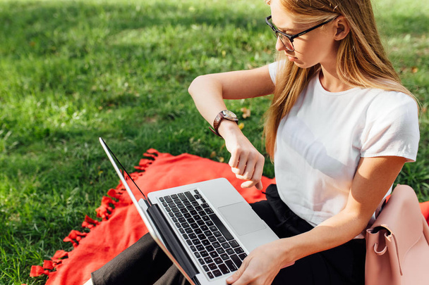 A imagem de uma bela menina estudante com óculos, que trabalha no laptop, e está atrasado olhando para o relógio, estar ao ar livre no Parque
 - Foto, Imagem