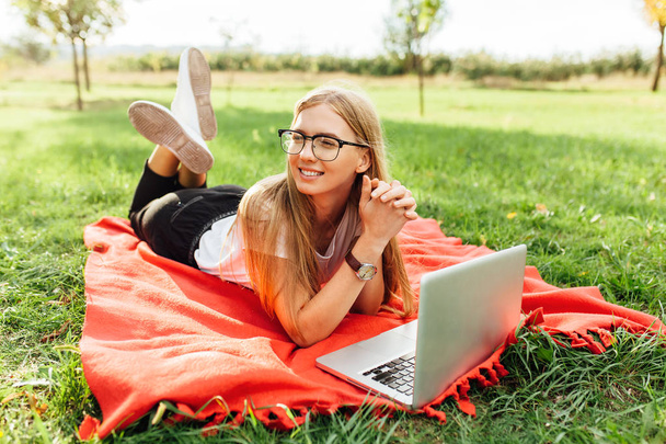 Изображение красивой студентки в очках, работающей на ноутбуке в хорошем настроении в парке, лежащей на одеяле
 - Фото, изображение