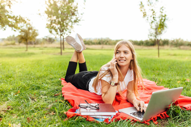 Image of a young beautiful student with glasses, talking on the phone, and sitting on a red blanket in the Park, with books and a laptop - Fotografie, Obrázek