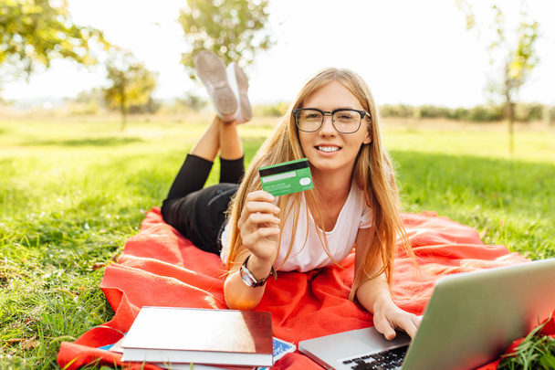 Beautiful girl in glasses with laptop touts a credit card sitting in the Park on the blanket - 写真・画像