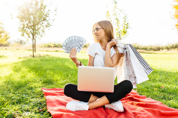 Beautiful business lady girl with glasses sitting on a blanket in the Park with a laptop and holding dollars in one hand and shopping in the other - Foto, immagini