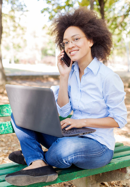 Dizüstü bilgisayarla çalışan ve telefon kullanan güzel melez bir kadın. Parkta bankta oturuyor, açık havada portre çiziyor, günlük kıyafetler ve gözlük takıyor.  - Fotoğraf, Görsel
