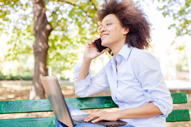 Laptop ile çalışma ve telefon kullanarak mutlu Afrikalı kadın. Park, açık portre, bankta oturmuş rahat kıyafetler ve gözlük takıyor  - Fotoğraf, Görsel