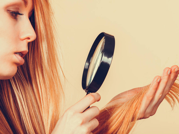 Haircare, health problem concept. Unhappy blonde woman looking at ends of her blonde hair through magnifying glass - 写真・画像