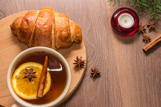 A cup of hot delicious tea with a croissant on a wooden platform with a New Year's decor in the form of Christmas tree branches of spices tinsel. - Photo, Image