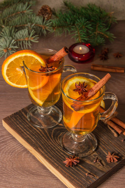 Dos vasos con delicioso té fragante de vino caliente con naranja y especias en una tabla de madera con ramas de un árbol de Navidad con una vela y oropel
. - Foto, Imagen