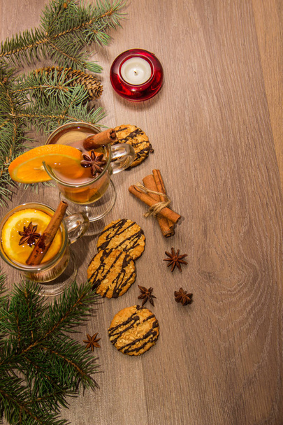 Dos vasos con delicioso té fragante de vino caliente con naranja y especias en una tabla de madera con ramas de un árbol de Navidad con una vela y oropel
. - Foto, Imagen