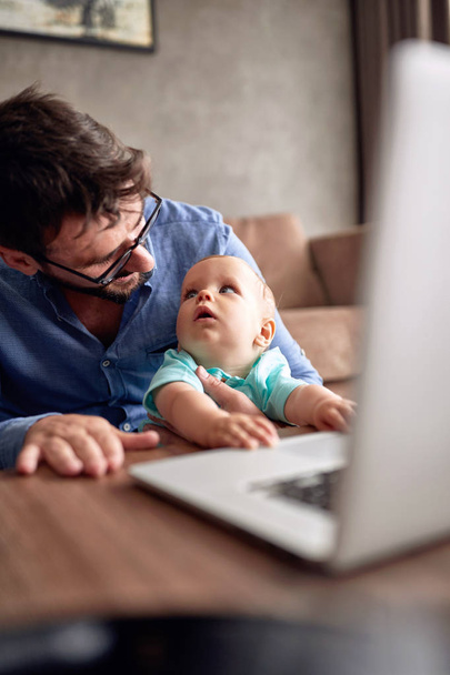 Young father using a laptop computer for work at home while looking after his baby son - Foto, Bild