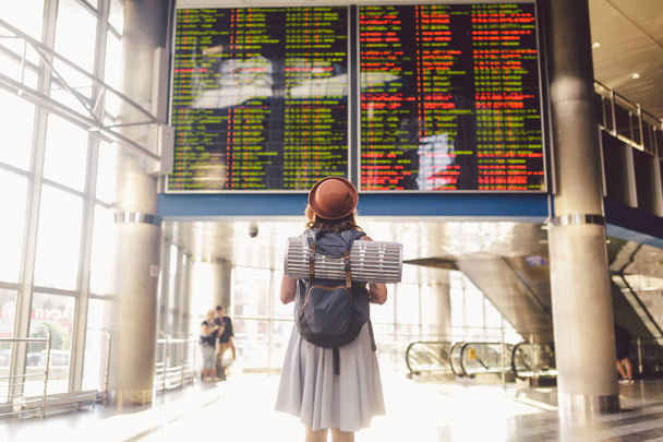Θέμα ταξιδιού δημόσιες μεταφορές. νεαρή γυναίκα στέκεται με την πλάτη στο φόρεμα και καπέλο πίσω από σακίδιο και Κατασκηνωτικός εξοπλισμός για τον ύπνο, πλών στρωμάτων μοιάζει χρονοδιάγραμμα πίνακα αποτελεσμάτων αεροδρόμιο σταθμό ηλιόλουστη ημέρα - Φωτογραφία, εικόνα