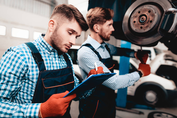 Young Mechanic Repairs Automotive Hub In Garage. Professional Uniform. Service Station Concept. Confident Engineer Stare. Detail Repairing. Under The Vehicle. Automobile Diagnostic. - Zdjęcie, obraz