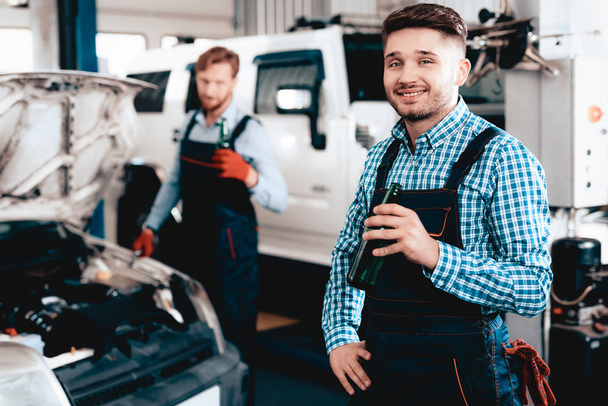 Two Young Mechanics Are Drinking Beer On A Service Station. Relaxing Concept. Having A Break Together. Bottle Of Alcohol. Resting Colleagues. Professional Uniform. Working In A Garage. - Foto, Imagen