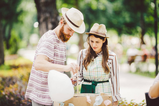Theme is a family small business cooking sweets. A pair of lovers A young man and woman trader in the hat the owner of the outlet makes candy floss, fairy floss or Cotton candy in the park in summer. - Photo, Image