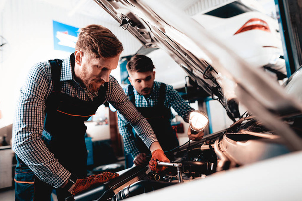 Young Auto Mechanics Repairs Car With A Wrench. Under The Hood. Professional Uniform. Using Flashlight. Engineering Specialist Team. Service Station. Automobile Diagnostic. Working In Garage. - Photo, image