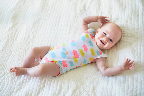 Baby girl lying on bed and smiling. Happy healthy little child laughing. Infant kid in sunny nursery - Photo, Image