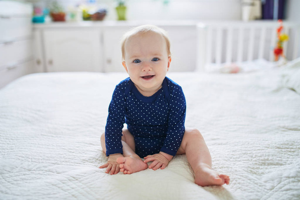 Een meisje dat op bed zit en lacht. Gelukkig gezond kind in blauwe kleren. Baby kind in zonnige kinderkamer - Foto, afbeelding