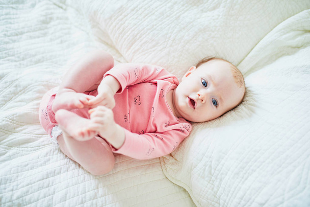 Baby girl lying on bed on her back and touching her toes. Happy healthy little child in pink clothes. Infant kid in sunny nursery - Foto, afbeelding