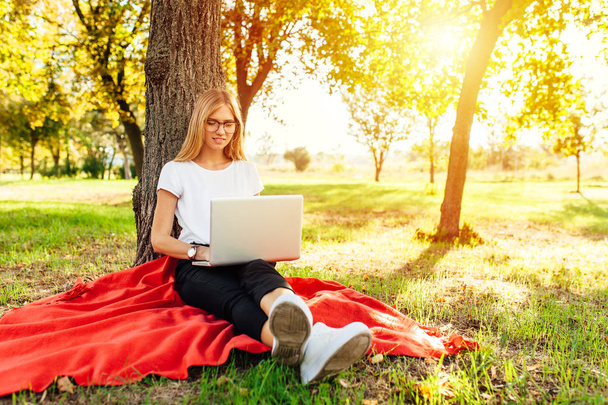 The image of a beautiful girl student with glasses, who works at the laptop, sitting on a red blanket in the Park near the tree, doing homework - Photo, image