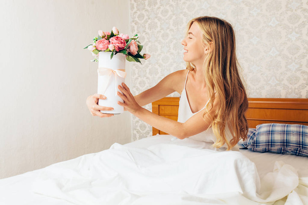 Young beautiful girl with peony flowers, sitting on the bed. She just woke up and was given a bouquet of flowers - Foto, Imagen