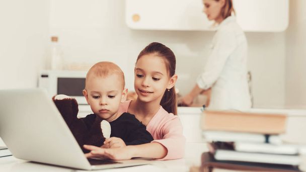 Young Girl Plays With Baby Brother On Kitchen. Happy Childhood Concept. Active Kids. Mom's Cooking Background. Family Holiday Leisure. Writing On The Laptop Keyboard. Parent's Duty. - 写真・画像