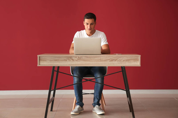 Νεαρός εργασίας με φορητό υπολογιστή ενώ κάθεται στο τραπέζι κατά χρώμα τοίχου - Φωτογραφία, εικόνα