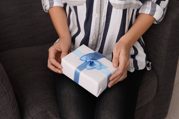 Woman holding gift box with blue ribbon while sitting in armchair - Photo, Image