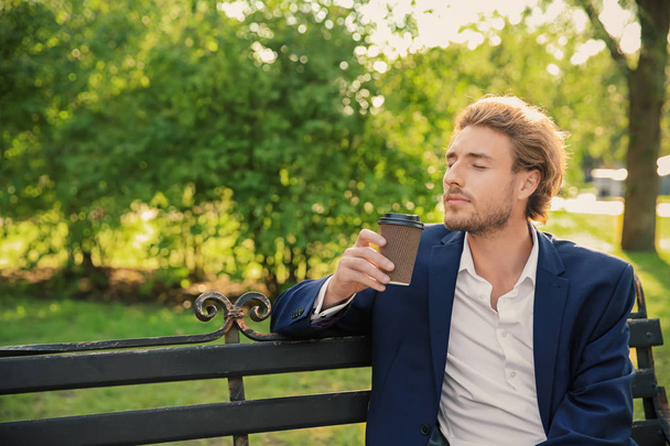 Красивый молодой бизнесмен с кофе отдыхает на скамейке в парке
 - Фото, изображение