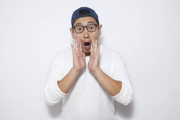 Photo image of young funny Asian man shocked or surprised expression with mouth open, touching cheek, while standing over white background - Foto, afbeelding