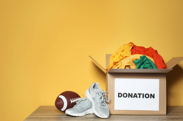Donation box with clothes, shoes and rugby ball on table against color background. Space for text - Фото, зображення